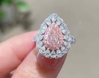 May include: A silver ring with a pear-shaped pink gemstone at its centre, encircled by smaller, clear stones. The ring's design is further enhanced by a halo of white stones. The overall aesthetic is one of elegance and sophistication.