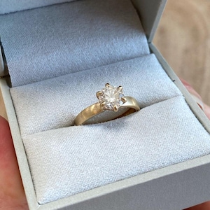 Può includere: Un anello di fidanzamento in oro con un diamante rotondo e chiaro in una scatola per anelli bianca.