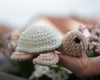 May include: A crocheted plush turtle toy with a light green shell and a light brown body. The turtle has black eyes and is being held in a hand.
