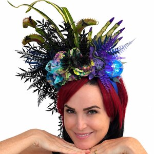 May include: A decorative headpiece featuring artificial Venus flytraps, black fern foliage, iridescent purple and blue flowers, a green skeletal hand, and a small blue orb. The headpiece is adorned with dark botanical elements and a spider accent.