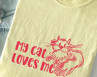 以下が含まれることがあります： 「My cat loves me」の赤い文字と赤い漫画の猫のイラストが描かれた、薄い黄色のTシャツ。 このシャツは柔らかい素材でできており、クルーネックです。