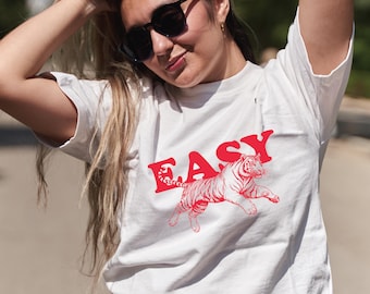 May include: White t-shirt with the word "EASY" in red, above a red tiger graphic. The tiger is depicted in a running pose. The shirt is a casual, everyday wear item.