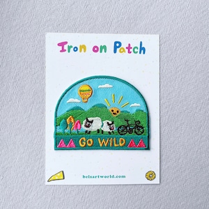 May include: Iron-on patch with a green, blue, and yellow landscape scene. The patch features two sheep, a bicycle, a hot air balloon, and a smiling sun. The text "GO WILD" is in yellow letters on a green background.