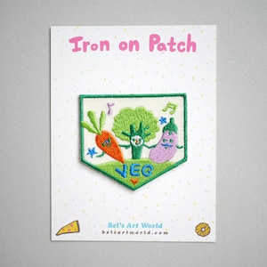 May include: Iron-on patch featuring a green broccoli, an orange carrot, and a purple eggplant, all with smiling faces. The patch is shaped like a green pennant with the word "VEG" embroidered in the center. The text "Iron on Patch" is printed in pink at the top of the image.