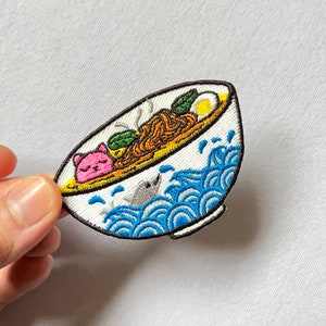 May include: A white bowl with blue and white waves on the outside, filled with ramen noodles, a pink pig, a green leaf, and a yellow egg.
