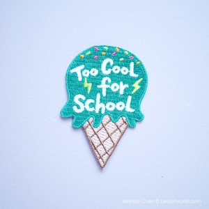 Peut inclure: Un cône de crème glacée bleu turquoise avec des pépites et le texte "Too Cool for School" en blanc avec des éclairs jaunes.
