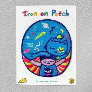 Könnte beinhalten: Bügeleisen-Patch mit einer blauen Katze mit einem Stern auf dem Kopf, die in einem blauen Kreis mit Sternen, Planeten und einem Kometen schläft. Die Katze ist von einem regenbogenfarbenen Heiligenschein umgeben. Der Text "Iron on Patch" ist über dem Patch gedruckt und "belsartworld.com" unter dem Patch.