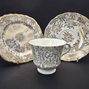 May include: A white teacup and two plates with a gold floral pattern. The teacup has a matching floral design and a gold rim. The plates have a scalloped edge. The set is on a black surface.
