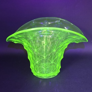 May include: A bright green glass vase with a flared, ruffled rim and a textured, geometric pattern. The vase has a wide, open top and a rounded base. The glass appears to glow, creating a striking visual effect. The vase is likely vintage.