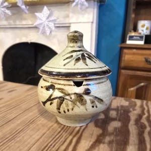 May include: A ceramic jar with a lid, featuring a beige and brown color scheme. The jar has a rounded body and a lid with a knob. The surface is decorated with a leaf-like pattern. The jar is sitting on a wooden table.
