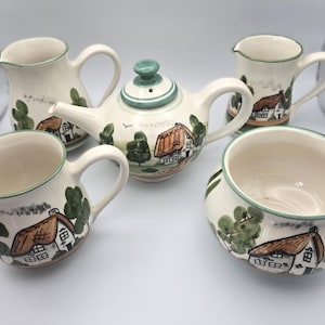 May include: A vintage ceramic tea set comprising a teapot, two mugs, a creamer jug, and a small bowl. Each piece is decorated with a hand-painted cottage scene in brown, green, and white. The set has a charming, traditional design.