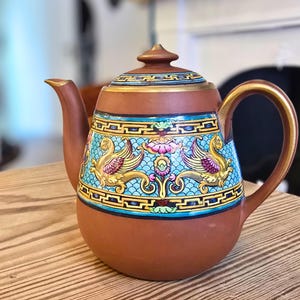 May include: A terracotta teapot featuring a decorative band with a bright blue background, gold Greek key borders, and colourful winged mythical creatures. The teapot has a matching lid with a gold rim, a curved spout, and a sturdy handle with gold accents.