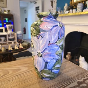 May include: A decorative ceramic vase with a floral design. The vase has a wide opening and is adorned with large, stylized flowers in blue, pink, and green on a white base. The vase is placed on a wooden table.