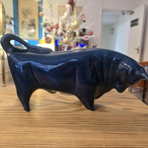 May include: A glossy, navy blue ceramic bull figurine. The sculpture has a curved tail and is posed in a charging stance. The bull is displayed on a light wood surface.