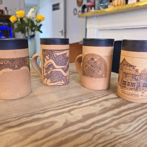 May include: Four handmade ceramic mugs with a tan base and dark blue rim. Each mug features a unique brown etched design, including figures, patterns, and a building with the word "TALGARTH". The mugs are on a wooden surface.