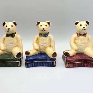 May include: Three ceramic teddy bear figurines. Each bear is seated on a decorative pillow. The bears have tan fur, brown paws, and wear bow ties in green, blue, and burgundy. The pillows are green, blue, and burgundy.