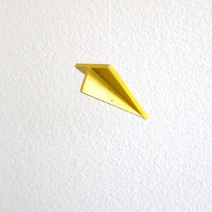 K&ouml;nnte beinhalten: Vertikales Bild mit drei Papierfliegern vor einem strukturierten wei&szlig;en Hintergrund. Der obere und untere Flieger sind wei&szlig;, der mittlere gelb. Sie h&auml;ngen an d&uuml;nnen F&auml;den, was einen spielerischen Effekt erzeugt.