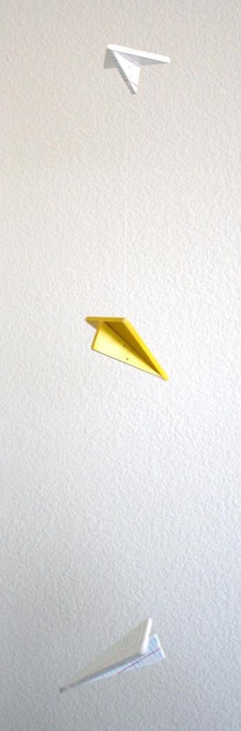 K&ouml;nnte beinhalten: Vertikales Bild mit drei Papierfliegern vor einem strukturierten wei&szlig;en Hintergrund. Der obere und untere Flieger sind wei&szlig;, der mittlere gelb. Sie h&auml;ngen an d&uuml;nnen F&auml;den, was einen spielerischen Effekt erzeugt.