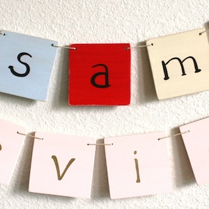 May include: A colorful banner with the name "Sammie" spelled out in black letters on individual wooden squares. The squares are strung together with twine and are in the colors red, blue, cream, pink, and green.