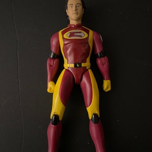 May include: A red and yellow action figure with a black belt and a large letter "E" on the chest. The figure has a black and yellow design on the arms and legs.