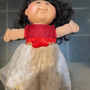 Può includere: Una bambola vintage con un viso rotondo, capelli neri e un top rosso. La bambola indossa una gonna bianca con pois dorati e un bordo dorato. Le braccia della bambola sono tese.