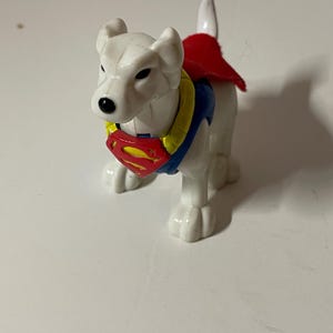 May include: White plastic toy dog wearing a red cape and a blue and yellow Superman symbol on its chest.