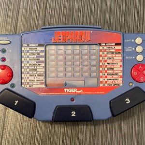 May include: A blue and black handheld electronic game with the word "JEOPARDY!" printed on the front. The game has a grid of squares with dollar amounts printed in each square. The game also has a list of categories on the left and right sides of the grid. The game is made by Tiger Electronics.