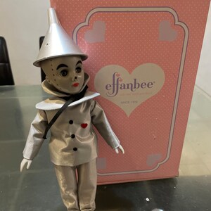 以下が含まれることがあります： オズの魔法使いのブリキ男に扮した銀色と白の磁器製のドール。ドールは銀色の帽子、銀色のジャケット、銀色のズボン、黒い靴を履いています。ドールは胸に赤いハートを持っています。ドールは、ピンクの箱の前にあるテーブルの上に立っています。箱には「effanbee」と「since 1910」の文字が印刷されています。