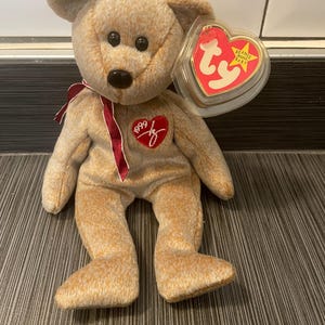 Puede incluir: Un oso de peluche Beanie Baby marrón y beige con una cinta roja alrededor del cuello. El oso tiene un corazón rojo en el pecho con el texto "1999" y una firma. El oso tiene una etiqueta en forma de corazón roja y dorada que dice "Beanie Babies Baby".