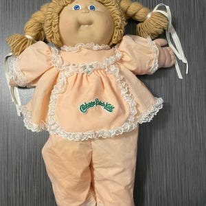 Puede incluir: Una muñeca Cabbage Patch Kids con pelo rubio de lana en trenzas, vestida con un vestido y pantalones de color melocotón con ribetes de encaje blanco. La muñeca tiene ojos azules y una sonrisa pintada. El vestido tiene un logotipo verde de Cabbage Patch Kids.