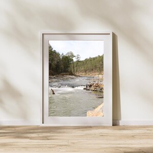 May include: A framed photograph of a river flowing through a forest. The water is white and foamy in some areas, and the trees are green and lush. The photo is in a white frame.