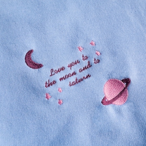 Pode incluir: Camisola azul claro com estrelas cor-de-rosa bordadas, uma lua crescente e um planeta com anéis. O texto "Love you to the moon and to saturn" está bordado em rosa.