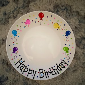 May include: White ceramic plate with a hand-painted design of colorful balloons and confetti. The plate says "Happy Birthday" in black lettering.