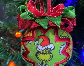 May include: A Christmas tree ornament featuring a green fabric ball with a Grinch character design. The ornament has red ribbon accents and a red and green bow at the top. The ornament is hanging on a Christmas tree.