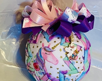 May include: A handmade ornament featuring a unicorn with a pink and purple color scheme. The ornament is made of fabric and has a white background with gold glitter stars. It is decorated with a purple and pink ribbon bow and a small unicorn charm.