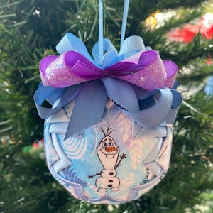 May include: A fabric Christmas ornament featuring Olaf from Frozen. The ornament is a light blue sphere with a printed design of leaves and snowflakes. It is topped with a multi-layered bow in shades of blue and purple, and a light blue ribbon for hanging.
