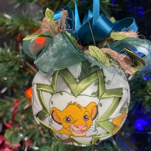 May include: A green and white fabric Christmas ornament with a picture of Simba from The Lion King. The ornament is decorated with a green ribbon and a brown twine bow.
