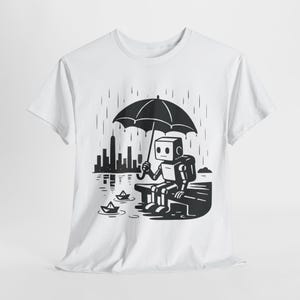 May include: White t-shirt featuring a black and white illustration of a robot sitting on a bench under an umbrella in the rain. The robot is looking at paper boats floating on the water, with a city skyline in the background.