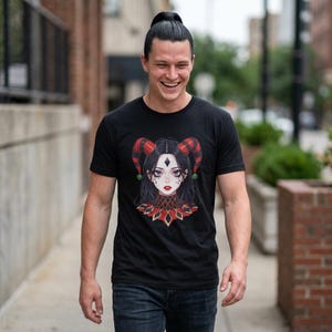 Może przedstawiać: Czarny t-shirt z krótkim rękawem z nadrukiem graficznym przedstawiającym postać z ciemnymi włosami, czerwono-czarnymi rogami błazna oraz pasującym czerwono-czarnym kołnierzem w romby na przodzie ubrania.