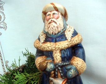 May include: A ceramic Santa Claus figurine, dressed in a navy blue coat with tan fur trim and matching hat. The figure holds silver bells with copper accents and wears blue mittens with snowflake designs. The figurine has a long white beard and is a festive holiday decoration.