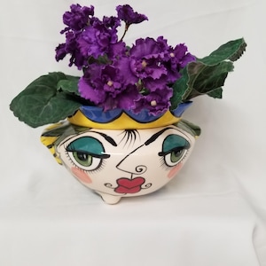 May include: A ceramic planter shaped like a human face with stylised features, including green eyes, red lips, and pink cheeks. The rim is painted yellow and blue, and the planter contains a vibrant purple African violet plant with green leaves.