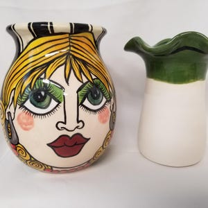 May include: Two ceramic vases. One is a round vase with a painted face design, featuring yellow hair, green eyes, and red lips. The other is a white vase with a green, ruffled top.