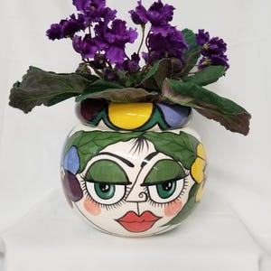 May include: A round ceramic planter featuring a hand-painted face with green eyes, long eyelashes, and red lips. The planter is decorated with green leaves and colorful flowers, and it contains a potted African violet plant with dark purple blooms.