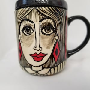 May include: A ceramic coffee mug with a black handle and a hand-painted design of a woman's face with red lips and earrings. The woman's hair is black and white, and her eyes are brown.