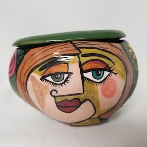 Peut inclure: Un pot en céramique en forme de visage avec un couvercle vert. Le visage a un design stylisé avec des cheveux orange, des yeux verts et bleus et un nez rose et jaune. Le pot est décoré de fleurs roses et jaunes.