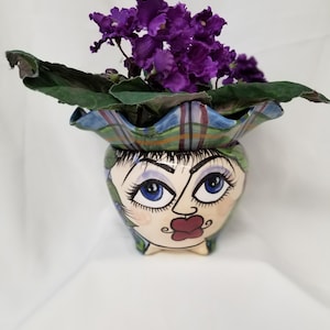 May include: A hand-painted ceramic planter featuring a stylized face with blue eyes and red lips. The planter holds a vibrant African violet with deep purple flowers and green leaves. The planter has a plaid rim.