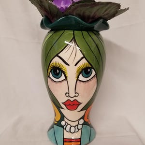 May include: A ceramic vase shaped like a human head with green hair, large blue eyes, long yellow eyelashes, and red lips. The vase features a white beaded necklace and a blue garment at the base. Purple flowers are placed inside the top opening.