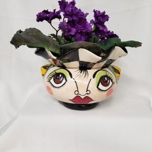 May include: A ceramic planter shaped like a human face with large green eyes, brown irises, and red lips. The rim features a black and white striped pattern. A vibrant purple African violet plant with dark green leaves sits inside the planter.