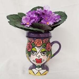 Peut inclure: Un mug en céramique violette avec une anse et un couvercle, orné d'un visage peint à la main avec des lèvres rouges, du fard à paupières vert et une couronne de roses roses. Le mug contient une violette africaine aux feuilles vert foncé.