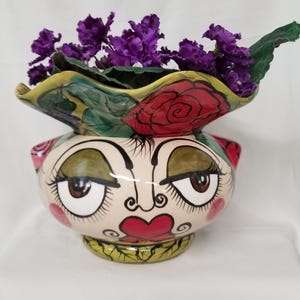 May include: A ceramic vase shaped like a stylized face with large brown eyes, green eyeshadow, and red heart-shaped lips. The rim features a ruffled edge painted with red roses and green leaves. Deep purple artificial flowers fill the vase.